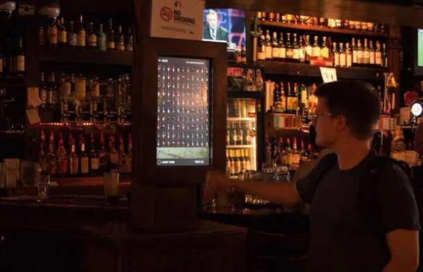 A customer at the bar at El Bait Shop, mid-selection, the wall of bottles behind him.